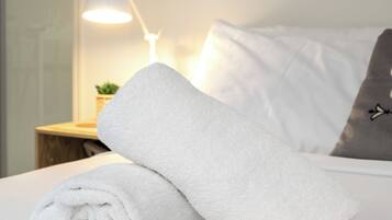 Una cama bien hecha con una toalla blanca enrollada, una lámpara de mesita de noche y una planta pequeña sobre una mesita de madera.