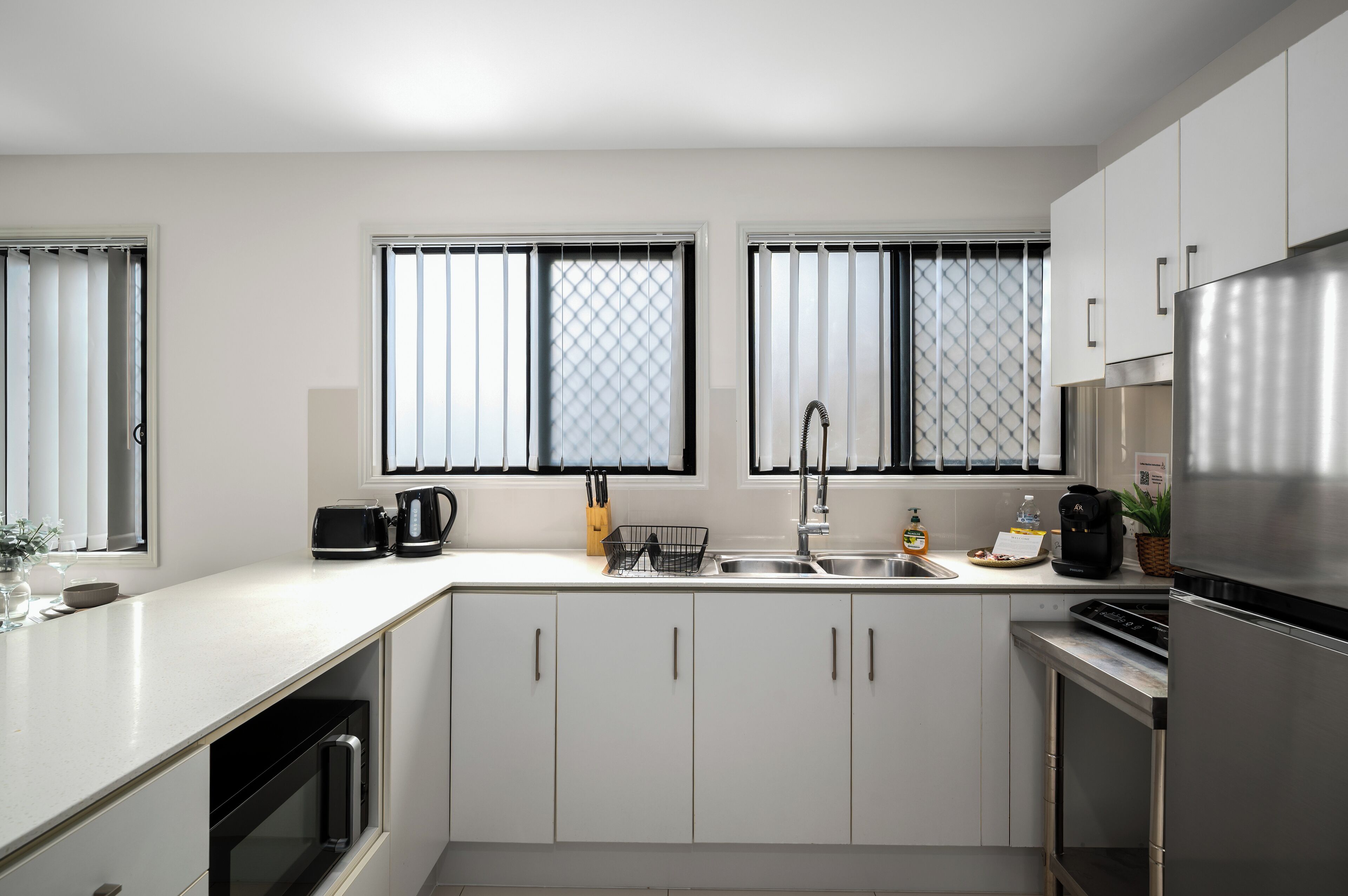 Une cuisine moderne avec des meubles blancs, un réfrigérateur en acier inoxydable, un évier et une fenêtre avec des stores.