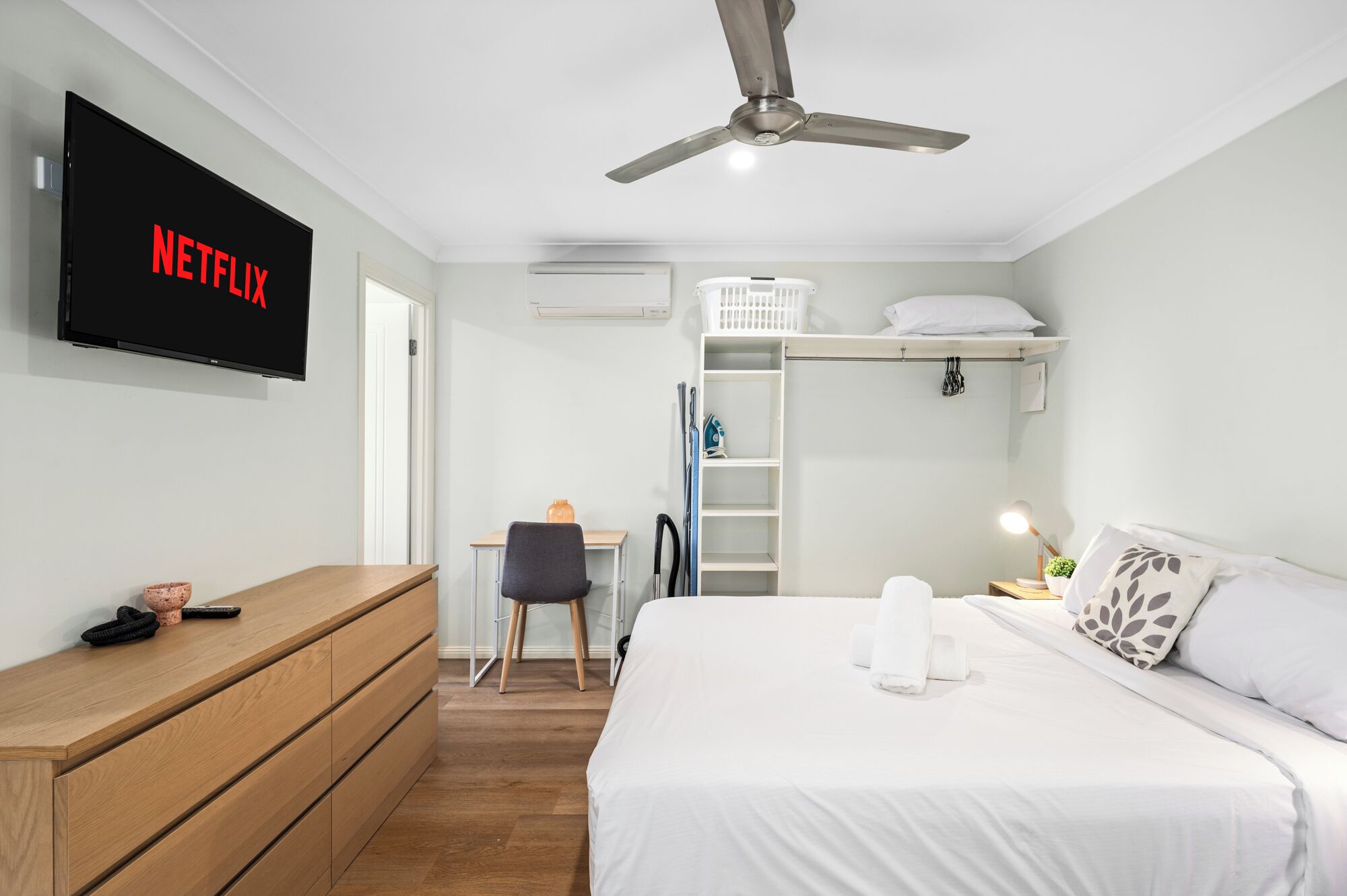A bedroom with a bed, a desk, a chair, a TV mounted on the wall, and a ceiling fan.