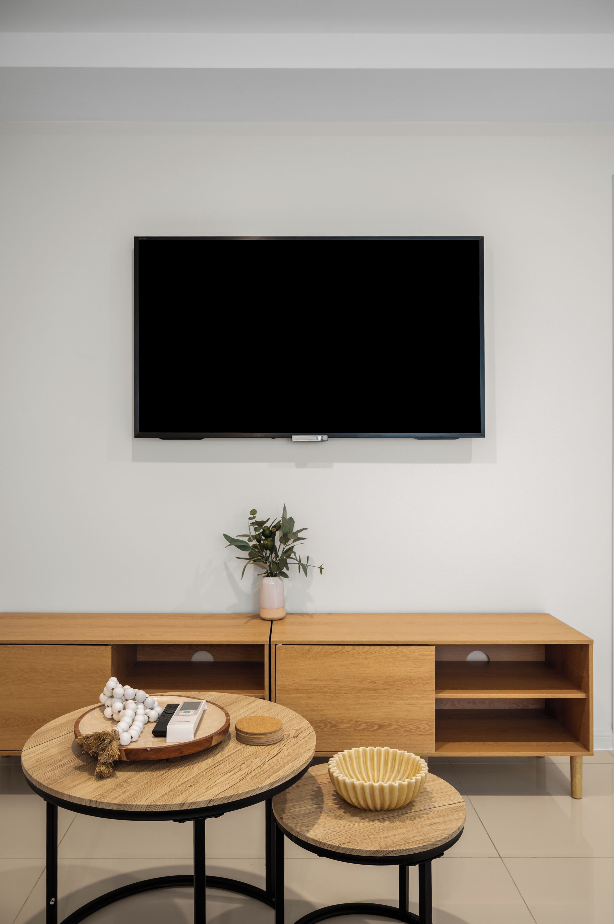A modern living room with a flat-screen TV mounted on the wall, a wooden console table with a potted plant, and two round tables with decorative items.