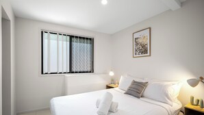 A neatly made bed with white linens and a rolled towel, a bedside lamp, a framed picture on the wall, and a window with blinds. - Smart Suites Self Check-in Apartments - 144 Dixon (Sunnybank)