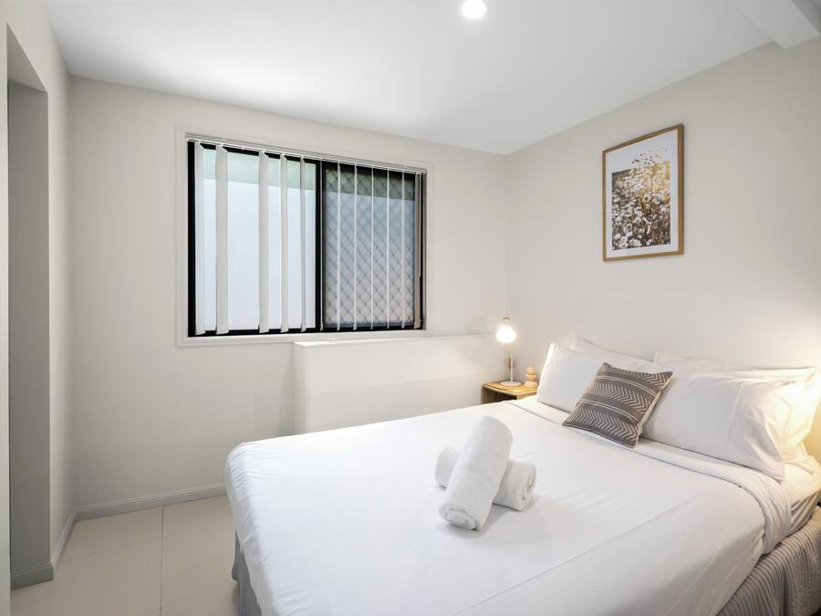 A neatly made bed with white linens and a rolled towel, a bedside lamp, a framed picture on the wall, and a window with blinds.