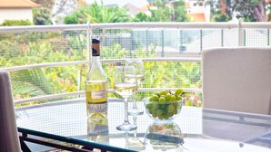 A table with a bottle of white wine, two wine glasses, and a bowl of green grapes on a balcony with a view of greenery and buildings. - Pino's Getaway by LovelyStay (Funchal)