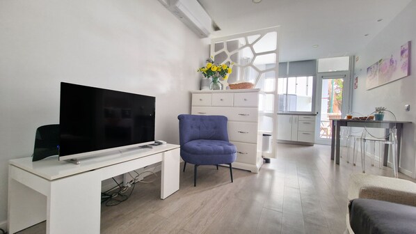 Uma sala de estar moderna com uma televisão de tela plana sobre uma mesa branca, uma poltrona azul e um aparador branco com um vaso de flores.