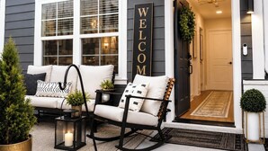 Exterior de una casa con un cartel que dice 'WELCOME', un porche con sofá y sillas, y una puerta que da al interior.