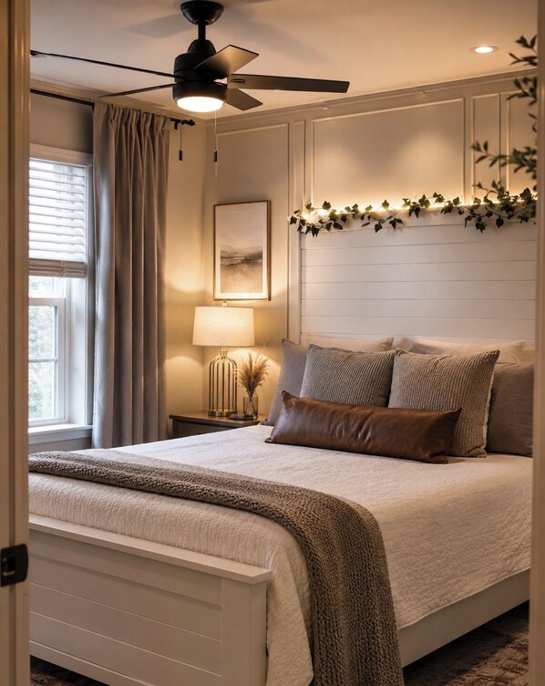 A bedroom with a bed, a ceiling fan, a nightstand with a lamp, and a window with curtains.