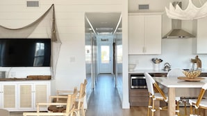 A spacious kitchen with white cabinetry, a dining area with wicker chairs, and a large TV mounted on the wall. - Cabell’s Coastal Cottage-Sleeps 10 Jockey’s Ridge 1 mile to beach Sunsets & Pets (Nags Head)