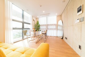A bright room with a yellow sofa, a dining table with chairs, and large windows with sheer curtains.