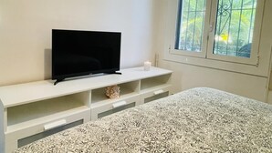 Un dormitorio con una televisión sobre un soporte blanco, una colcha estampada y una ventana con vistas a zonas verdes.