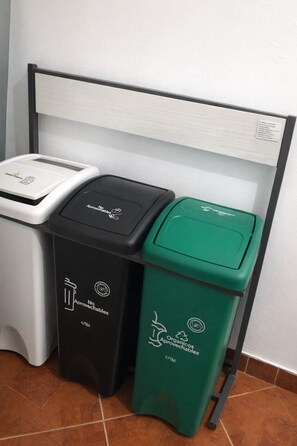 Drie vuilnisbakken met recycling symbolen, één zwart, één wit en één groen, geplaatst tegen een muur.