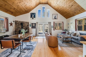 Een ruime loft met een hoge houten balkenplafond, een stenen openhaard, een eethoek met een lange tafel en stoelen, een gezellige zithoek met een bank en fauteuils, en grote ramen met uitzicht op de omgeving.