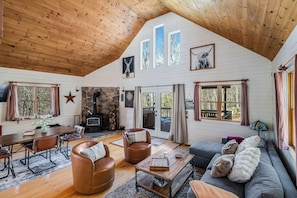 Een ruime woonkamer met een houten plafond, een stenen open haard en een groot raam met uitzicht op een terras.