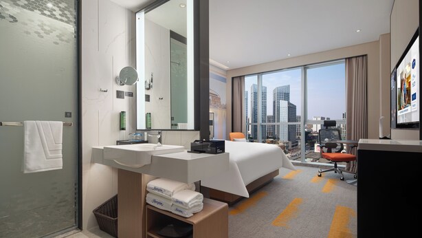 Una habitación de hotel moderna con una cama grande, dos lavamanos, un espejo amplio, un escritorio y vistas al horizonte de la ciudad.