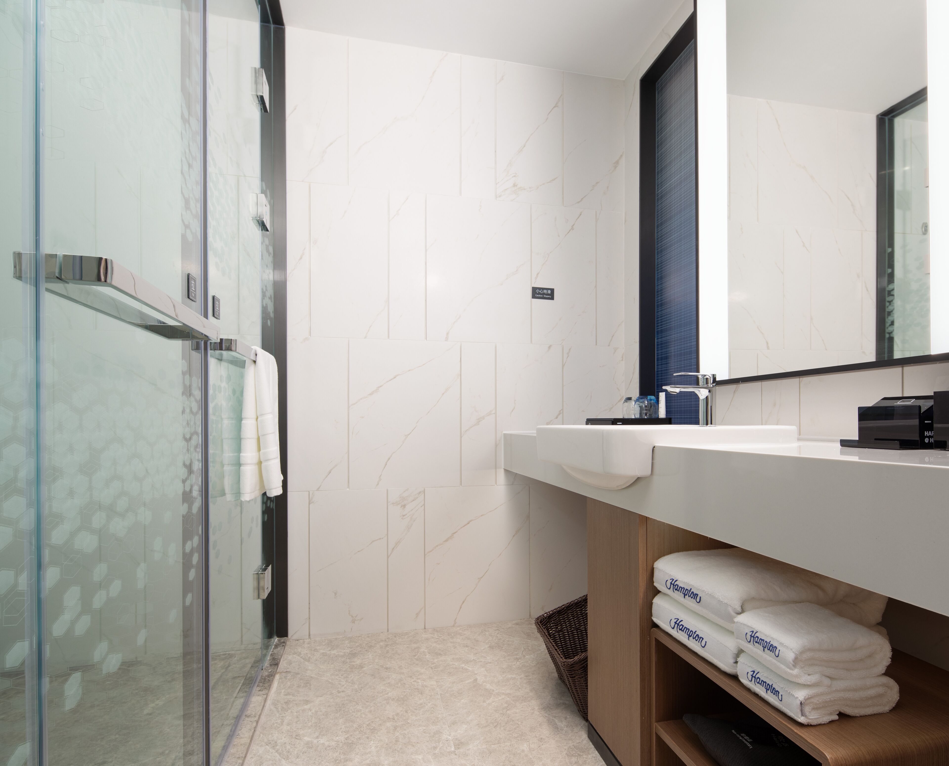 A modern bathroom with a glass shower enclosure, white marble tiles, a wooden vanity with a mirror, and neatly folded towels.