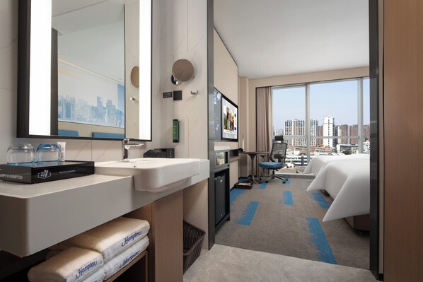 A hotel room with a large window, a bed, a desk, a chair, a sink, and a mirror. - HamptonHiltonChengduRailwayNorthStation (Chengdu)