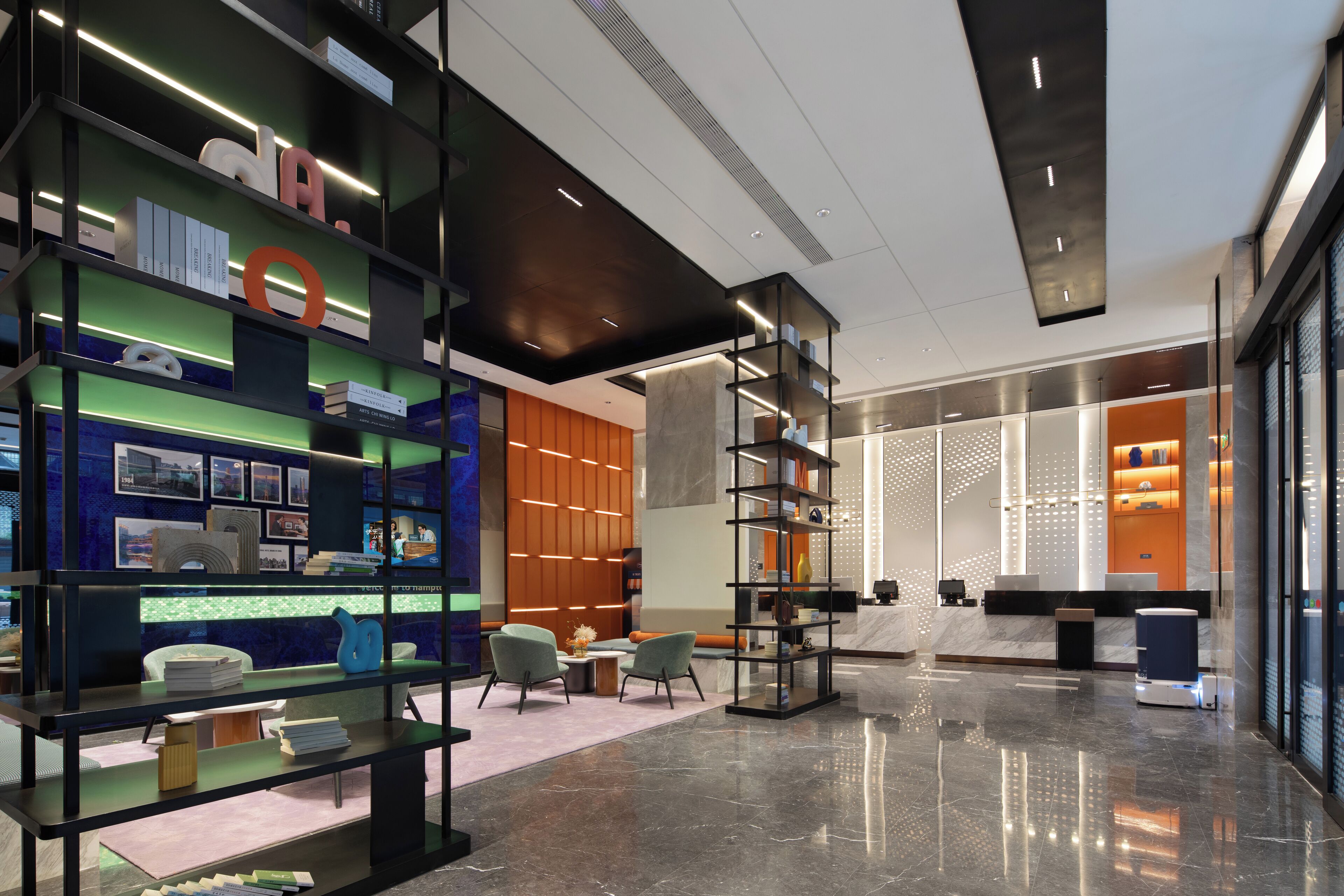 A modern lobby with a glass reception desk, a seating area with two chairs, and a bookshelf with various items.