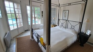 Een hemelbed met een witte spreideken, een nachtkastje met een lamp, en uitzicht op het landschap door een deur.