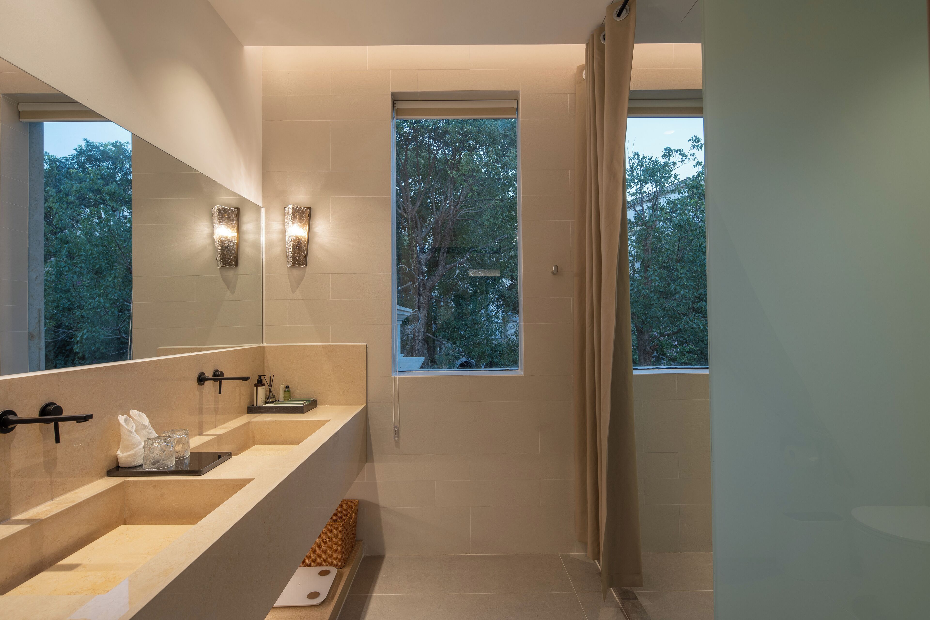 A modern bathroom with a large mirror, two wall sconces, a window, and a shower area.