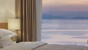 A hotel room with a bed, a nightstand with a lamp, and a view of the sea and mountains. - Miyue Roman Holiday Bubble Pool hotel (Shenzhen)