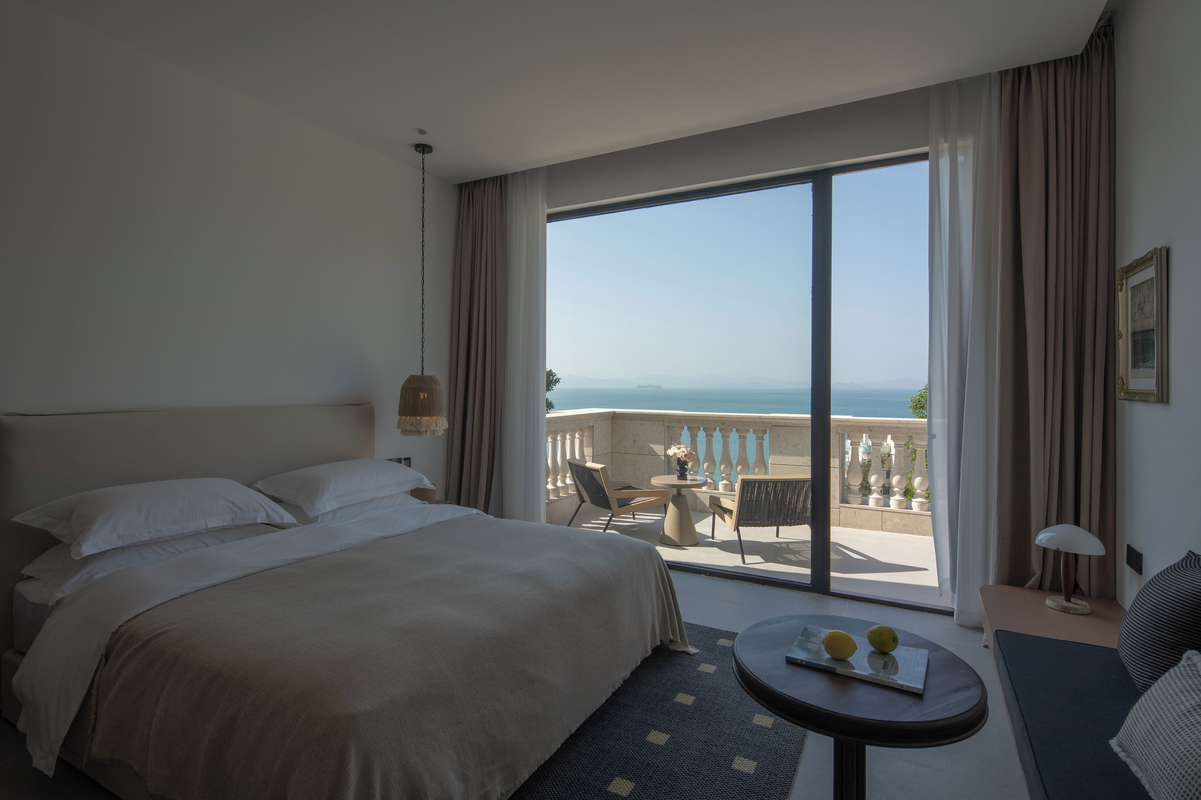 Une chambre d’hôtel avec un grand lit, une petite table ronde sur laquelle sont posés deux citrons, un canapé et un balcon offrant une vue sur la mer.