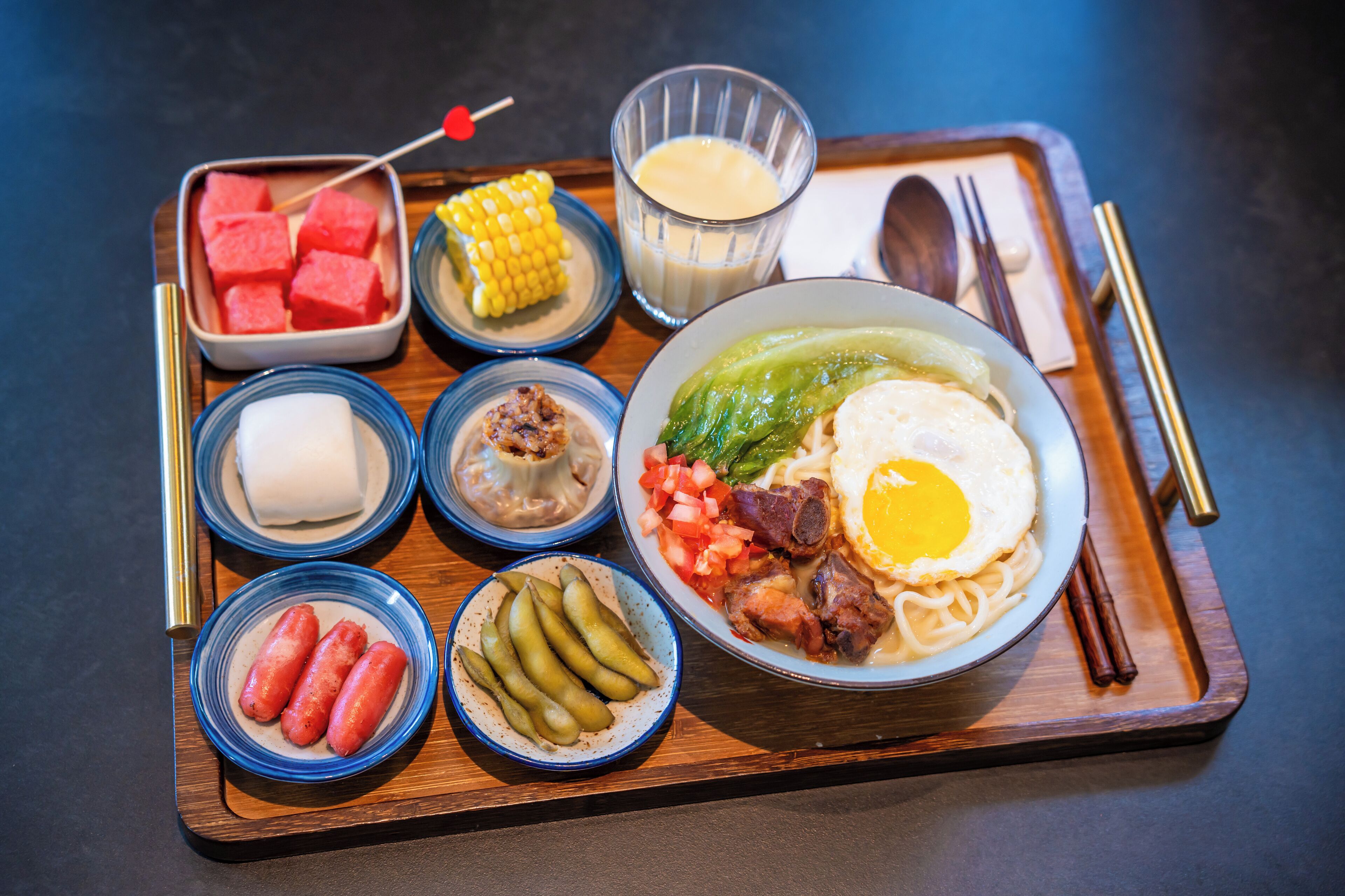 Una bandeja con diversos alimentos, incluyendo un tazón de fideos con verduras y carne, un vaso de leche, elote, cubos de sandía, pepinillos y salchichas.