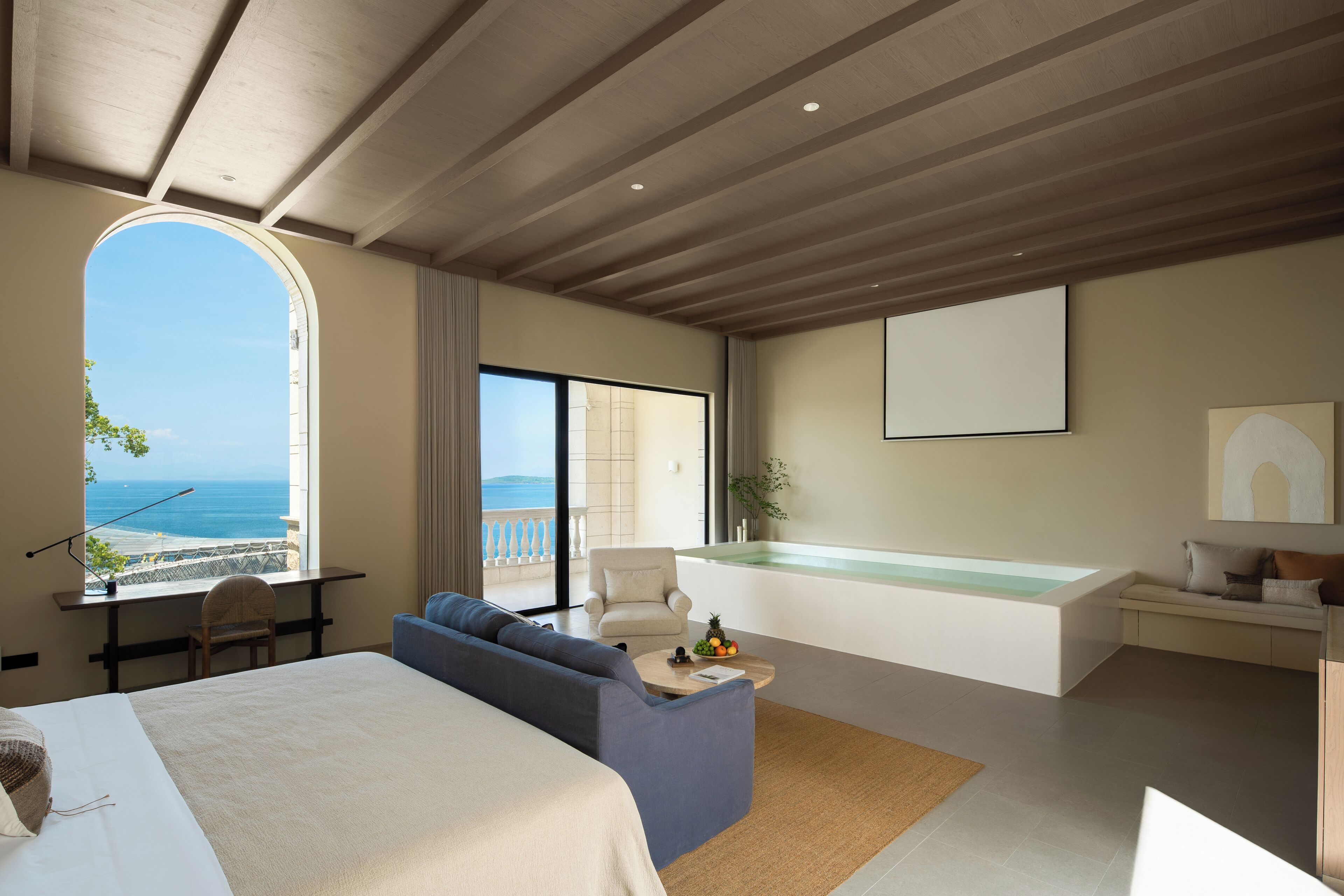 A modern hotel room with a large bed, a sofa, a desk, a balcony with a view of the sea, and a jacuzzi.