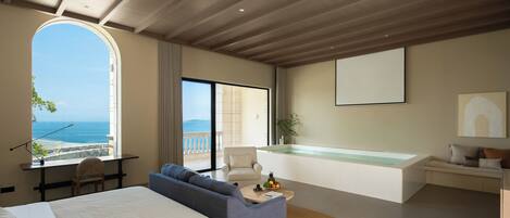 A modern hotel room with a large bed, a sofa, a desk, a balcony with a view of the sea, and a jacuzzi.