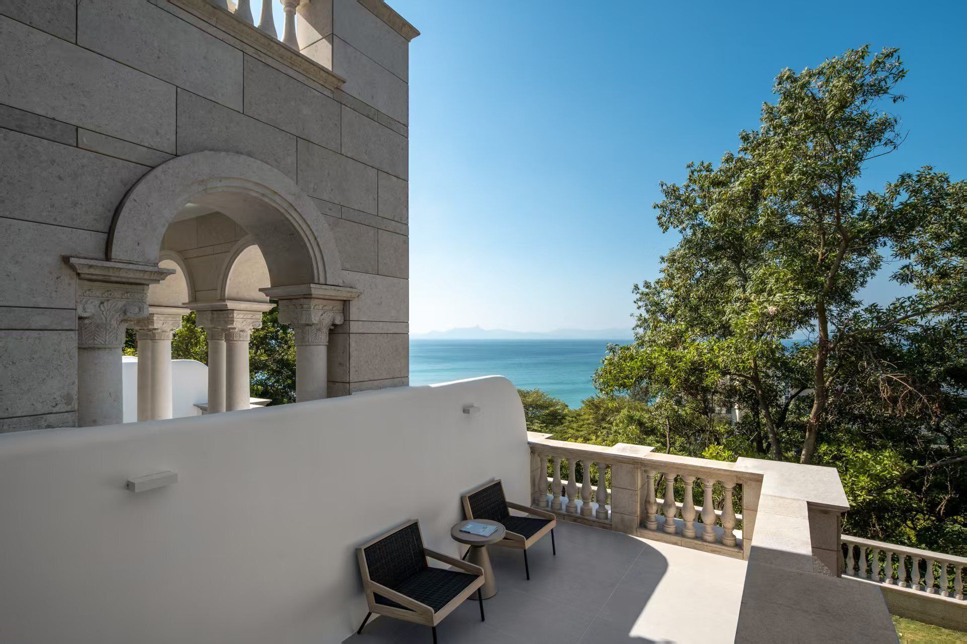 A balcony with two chairs and a table, overlooking a scenic view of the sea and a mountainous horizon.
