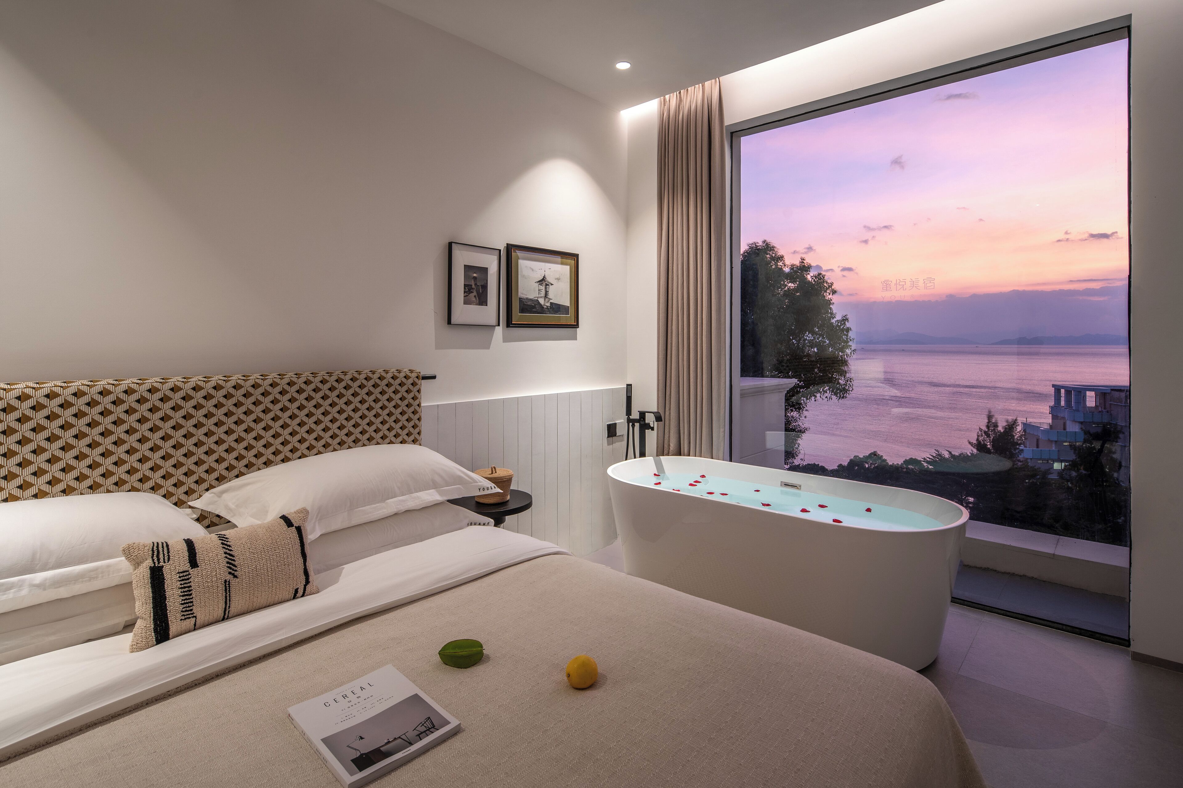 A modern hotel room with a large window, a bathtub, and a bed with a patterned headboard.