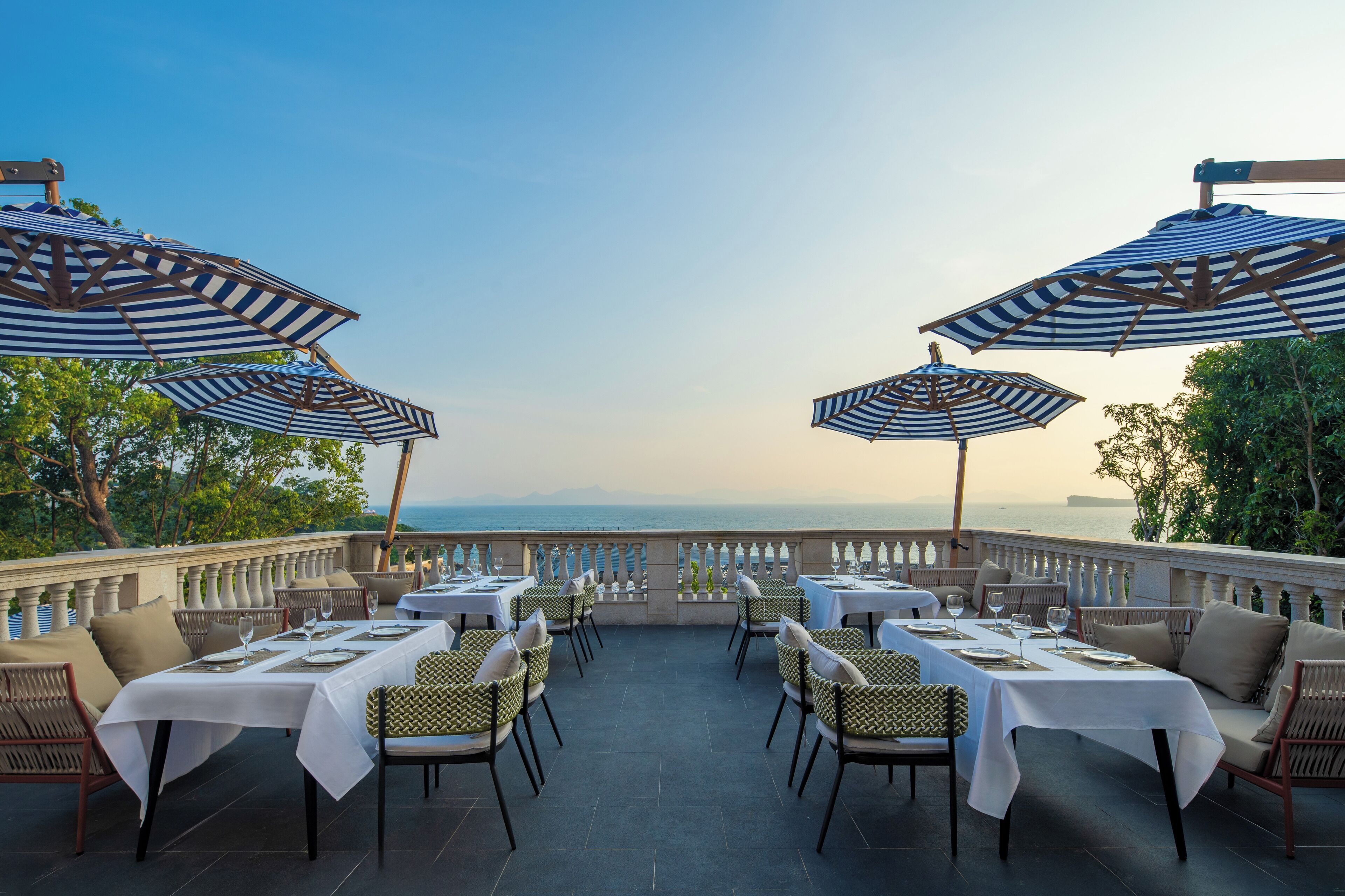 An outdoor dining area with tables and chairs, striped umbrellas, and a view of the sea.