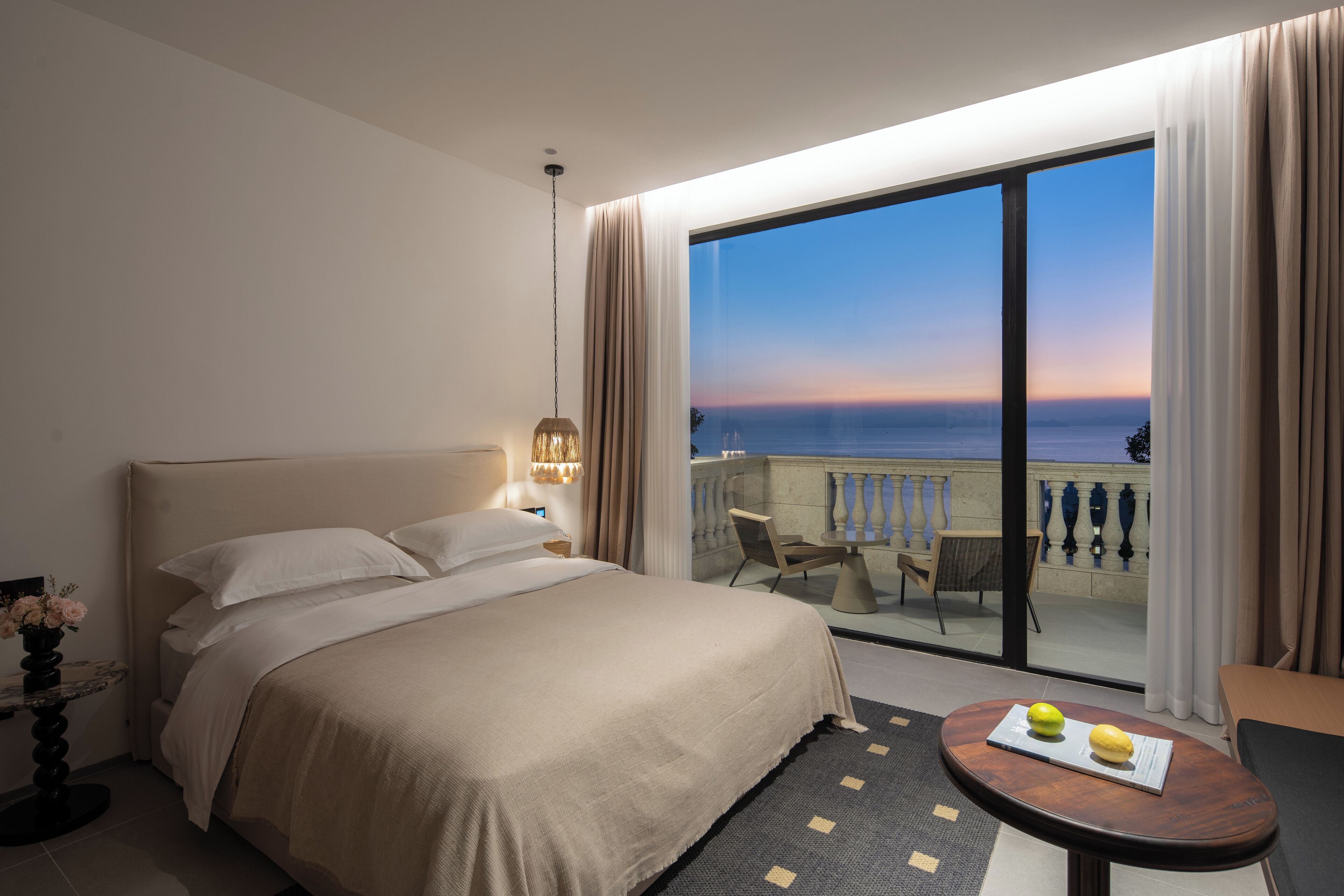 Habitación de hotel con una cama grande, una pequeña mesa con fruta, un balcón con vistas al mar y un comedor con sillas.
