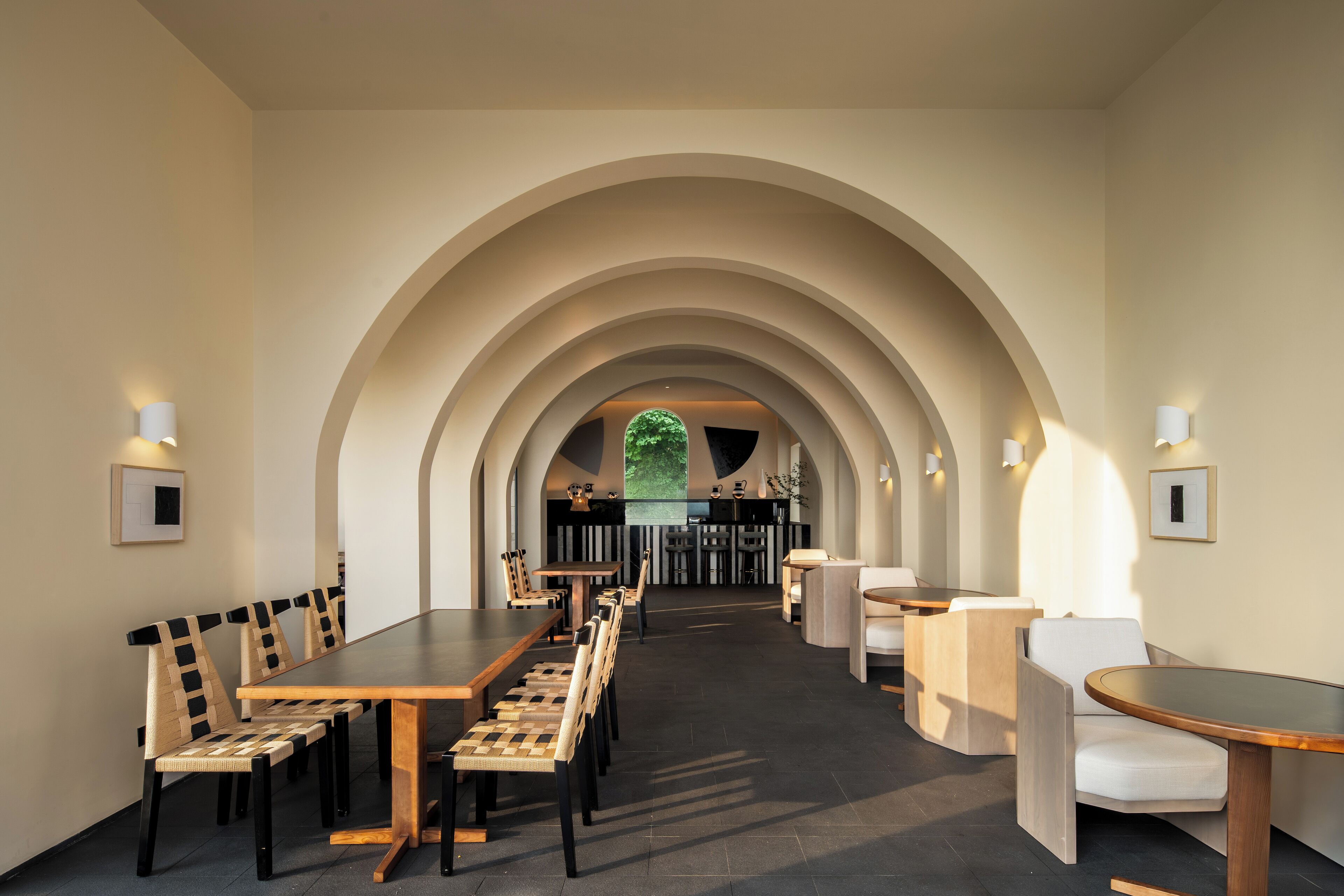 A modern dining area with a long table and chairs, an arched ceiling, and a view of a bar area in the background.