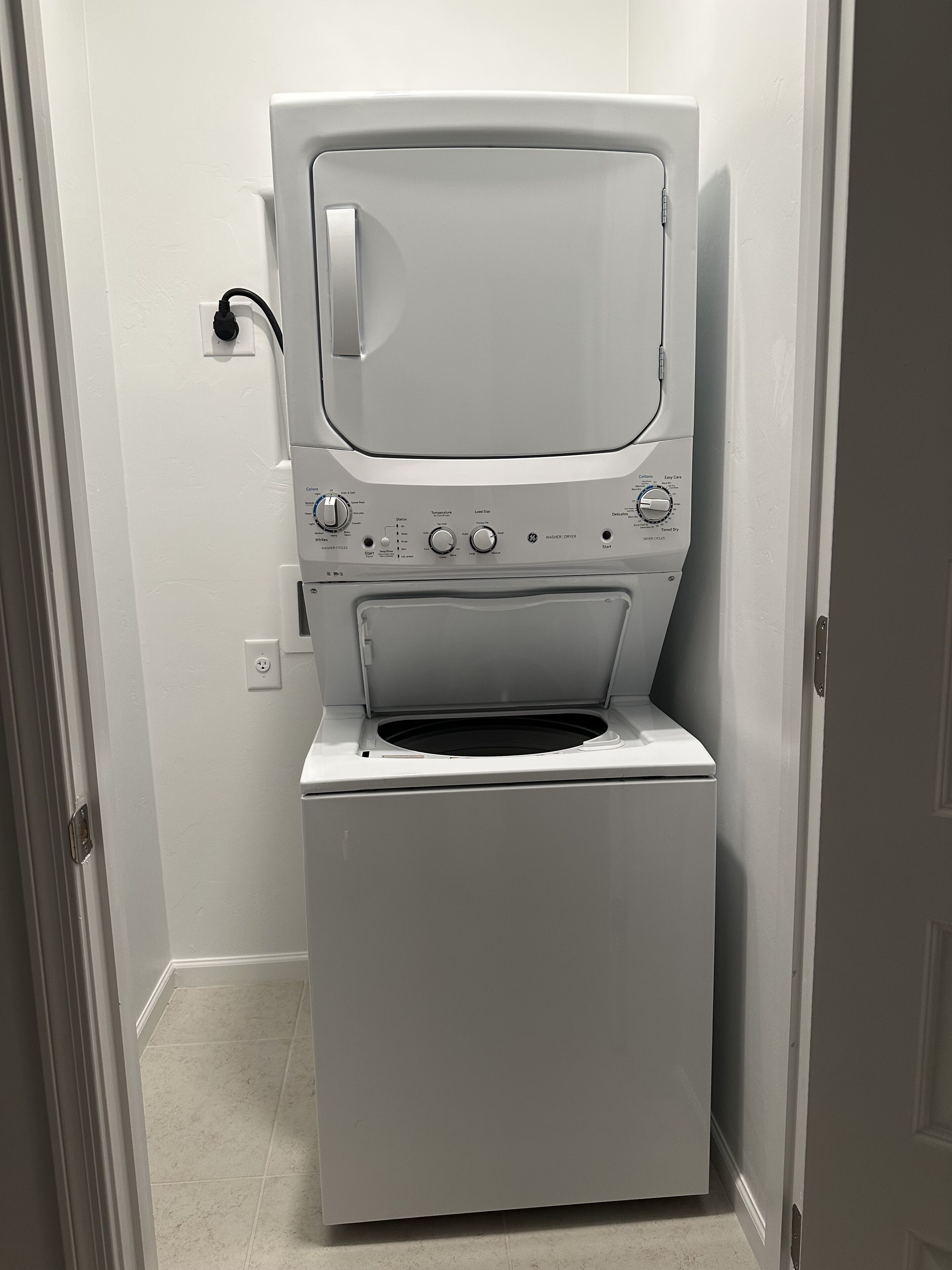 A compact, white, front-loading washing machine in a small laundry room with a door on the left and a wall-mounted electrical outlet.