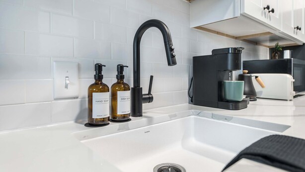 Una cocina moderna con un lavamanos blanco, grifo negro y una máquina de café. Hay dos botellas de detergente para lavar platos sobre la encimera.