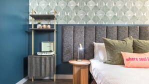 Un dormitorio con cama, mesita de noche con lámpara, estante con adornos y un papel tapiz estampado.