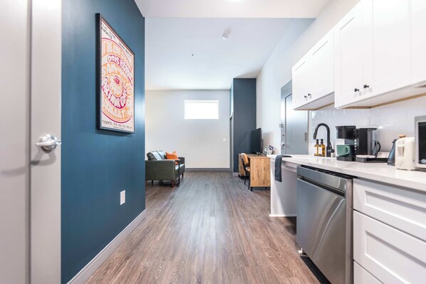 A modern kitchen with white cabinets, a dishwasher, and a dining area with a wooden table and chairs. - ADA Studio Suite w/ Walk Score 72 & Near Cherry St (Macon)