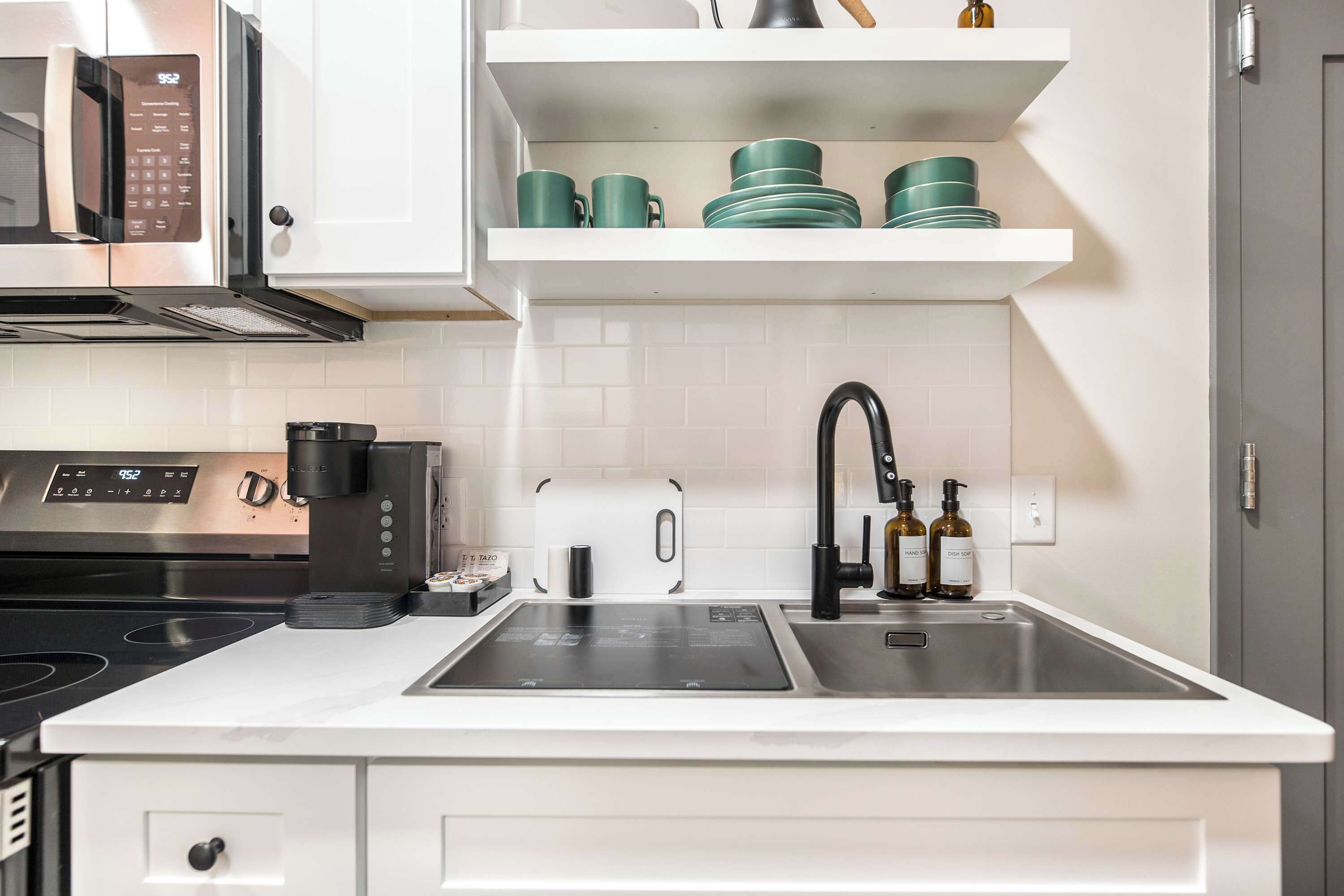 Een moderne keuken met een ingebouwde magnetron, fornuis en koffiezetapparaat. Er is een spoelbak met kraan en een plank erboven met groene servies.