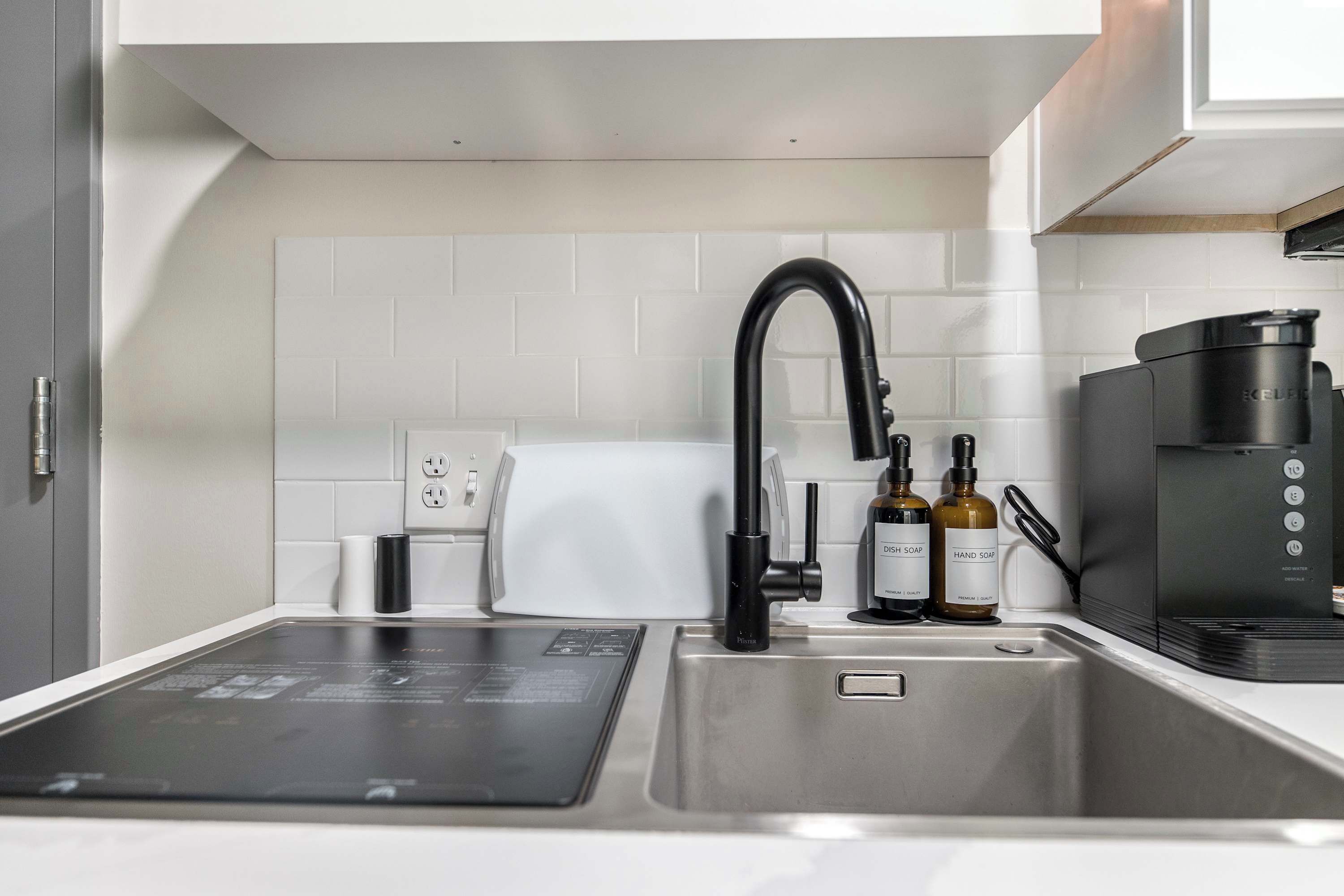 Una cocina moderna con un lavamanos de acero inoxidable, un grifo negro, una cafetera y dos botellas de jabón para manos.
