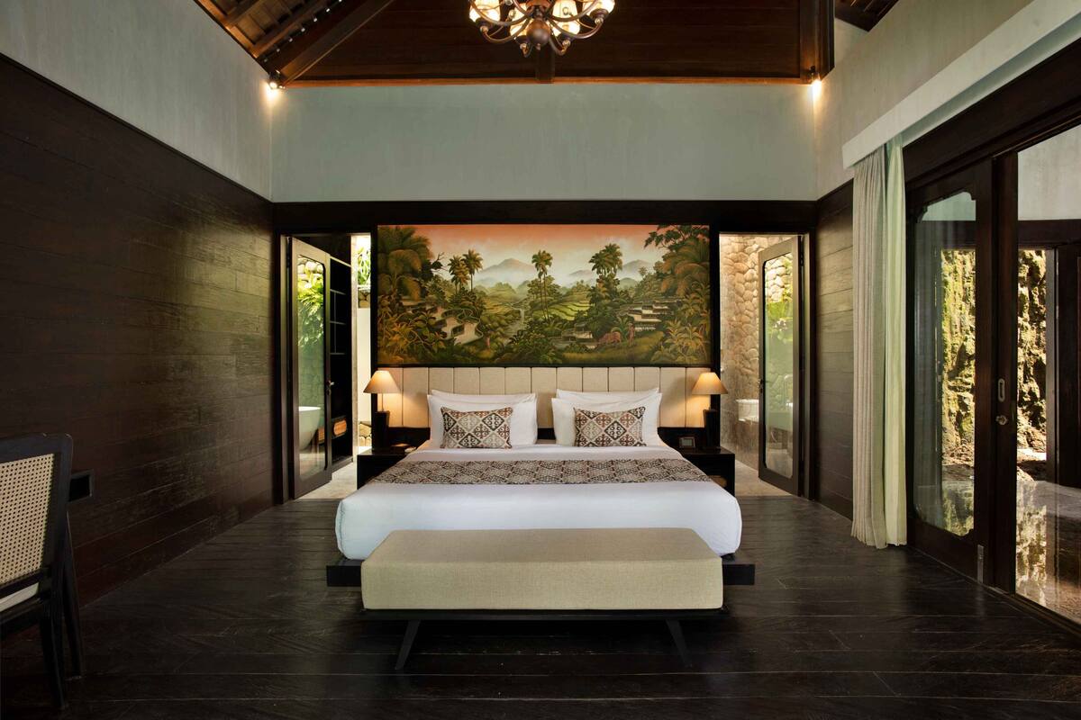 A large bed with a headboard, bedside lamps, and a view of a landscape painting.