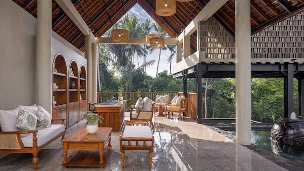 A spacious indoor area with a high ceiling, wooden furniture, and a view of palm trees outside. - Adiwana Unagi Riverfront (Mas)