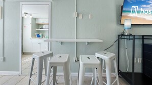 Ένα δωμάτιο με μικρή κουζίνα, μια τηλεόραση τοποθετημένη στον τοίχο και ένα γραφείο με καρέκλα.