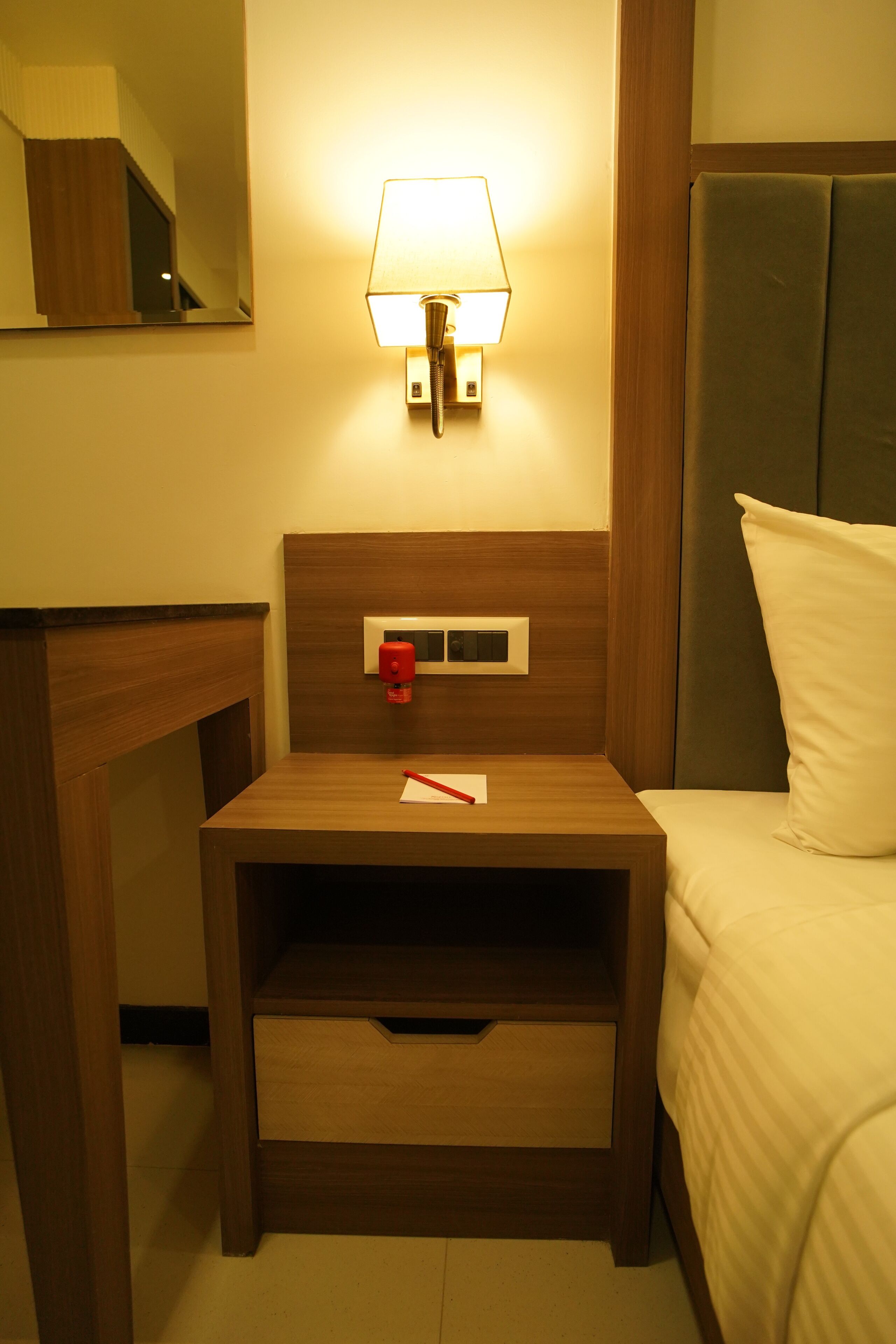 A hotel room with a wooden nightstand, a bedside table, a lamp, a bed with white bedding, and a mirror on the wall.