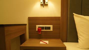 A hotel room with a wooden nightstand, a bedside table, a lamp, a bed with white bedding, and a mirror on the wall.