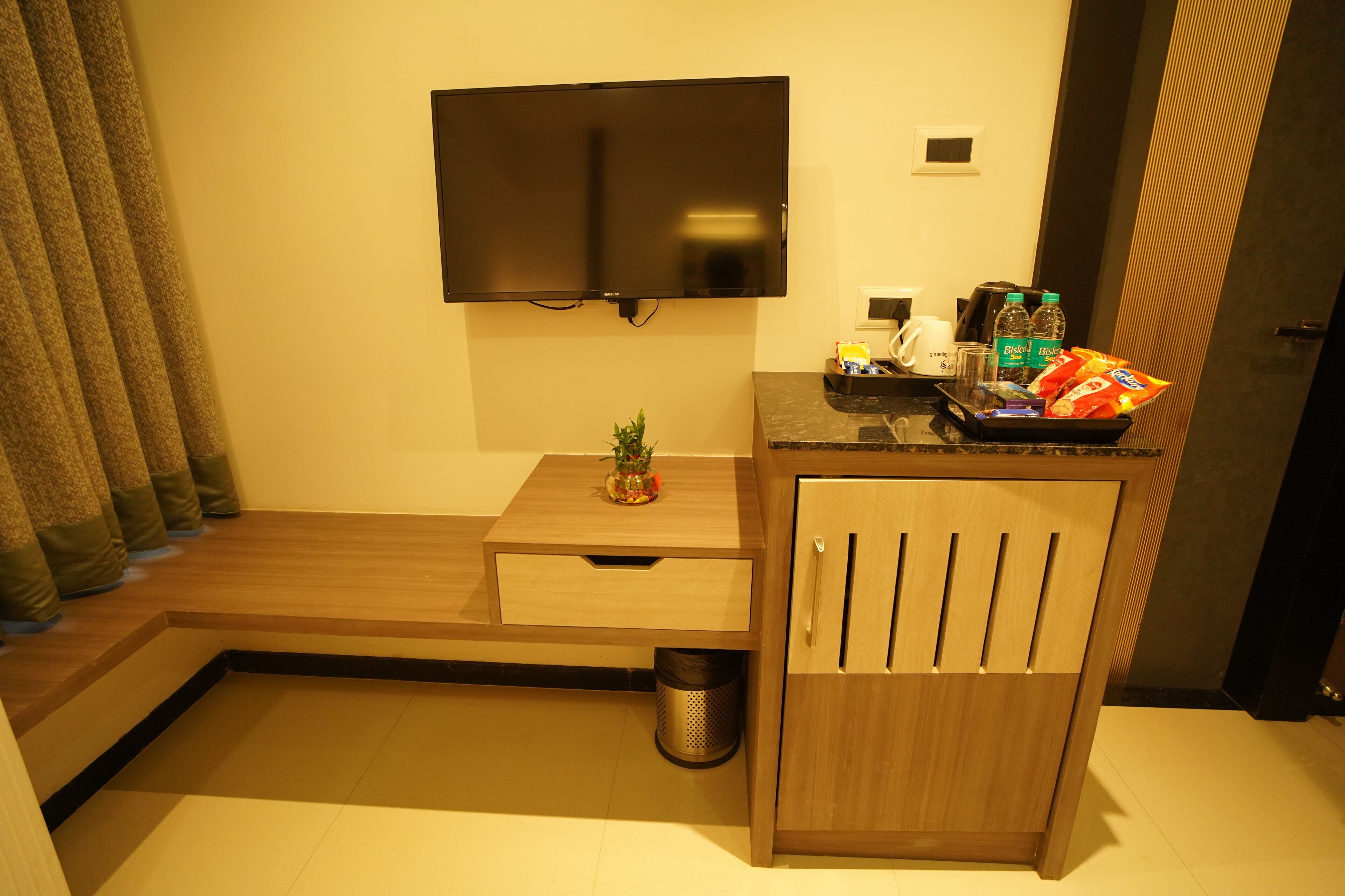 A hotel room with a flat-screen TV mounted on the wall, a small wooden table with a plant, a trash can, and a minibar with snacks and drinks.