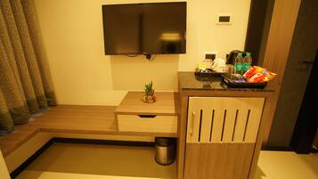 A hotel room with a flat-screen TV mounted on the wall, a small wooden table with a plant, a trash can, and a minibar with snacks and drinks.