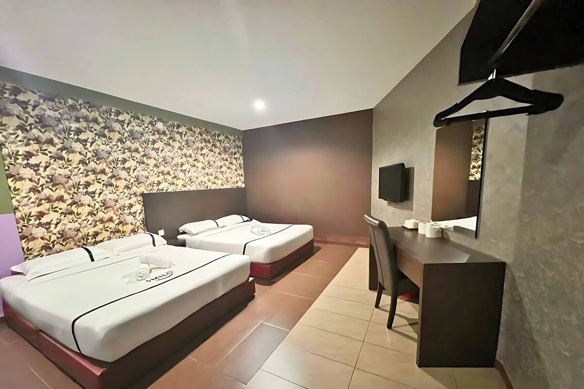 Ein Hotelzimmer mit zwei Betten, einem Schreibtisch, einem Sessel, einem Fernseher und einer Wand mit floralem Muster.