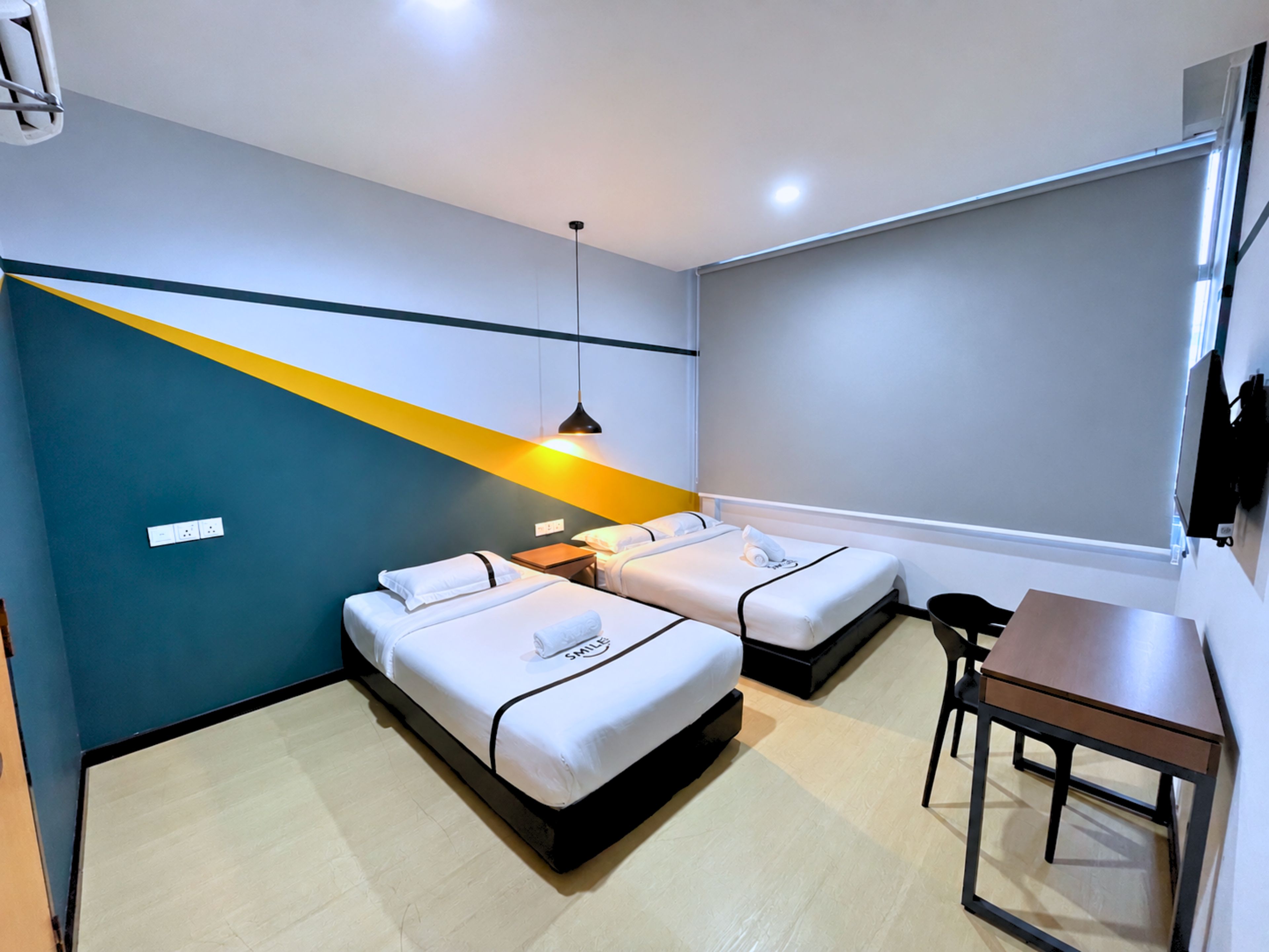 A hotel room with two beds, a desk, a chair, and a modern pendant light.