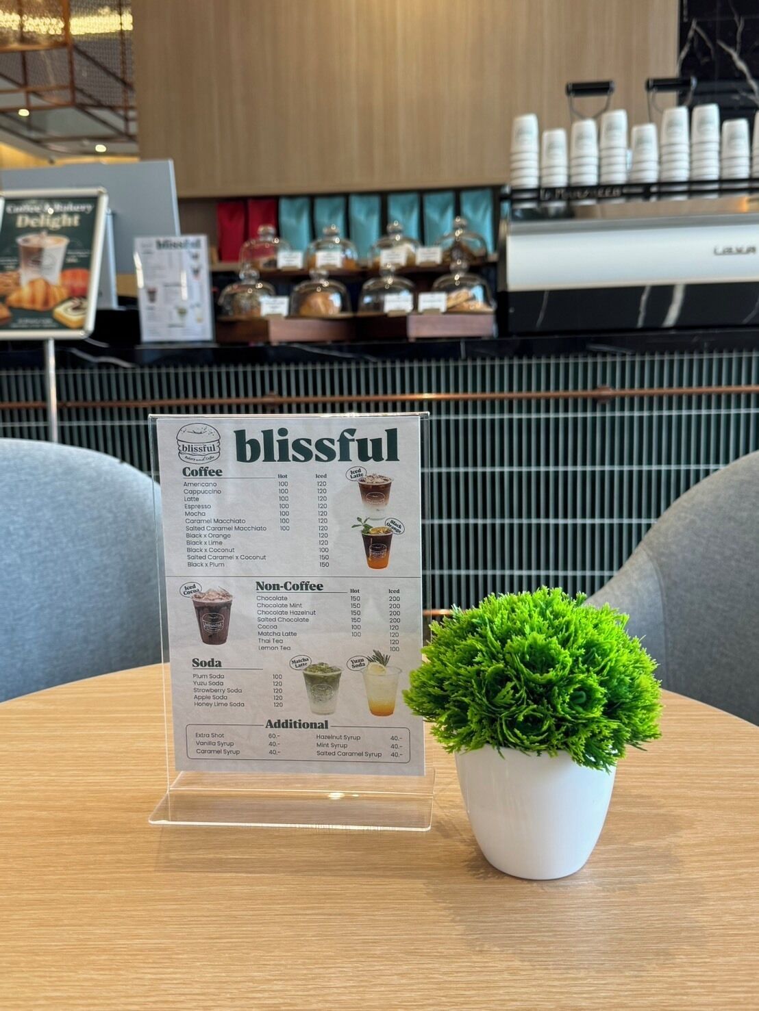 A menu board titled 'blissful' with coffee, non-coffee, and soda options, placed on a table with a potted plant.