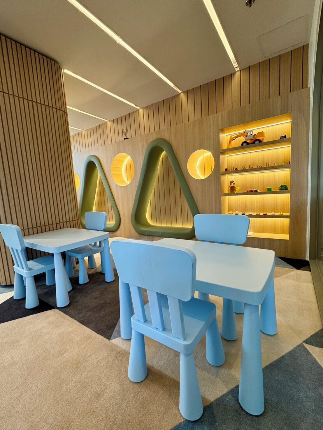 A children's play area with tables and chairs, a wooden wall with shelves, and a ceiling with recessed lighting.