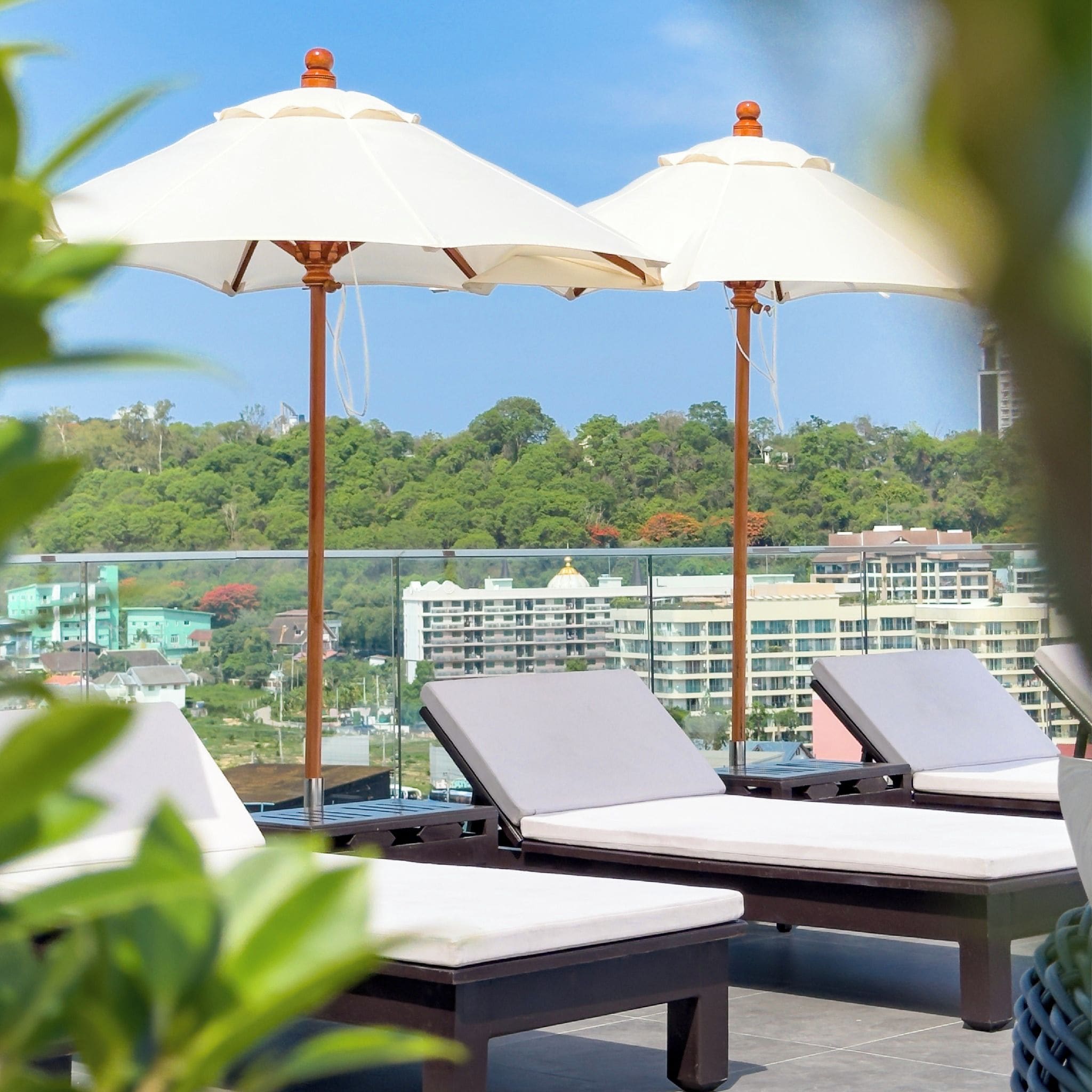 A rooftop lounge area with two sunbeds and umbrellas, overlooking a cityscape and greenery.